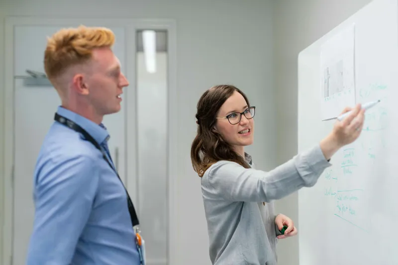Woman and man finding a root cause using five whys in front of a whiteboard in conference room.