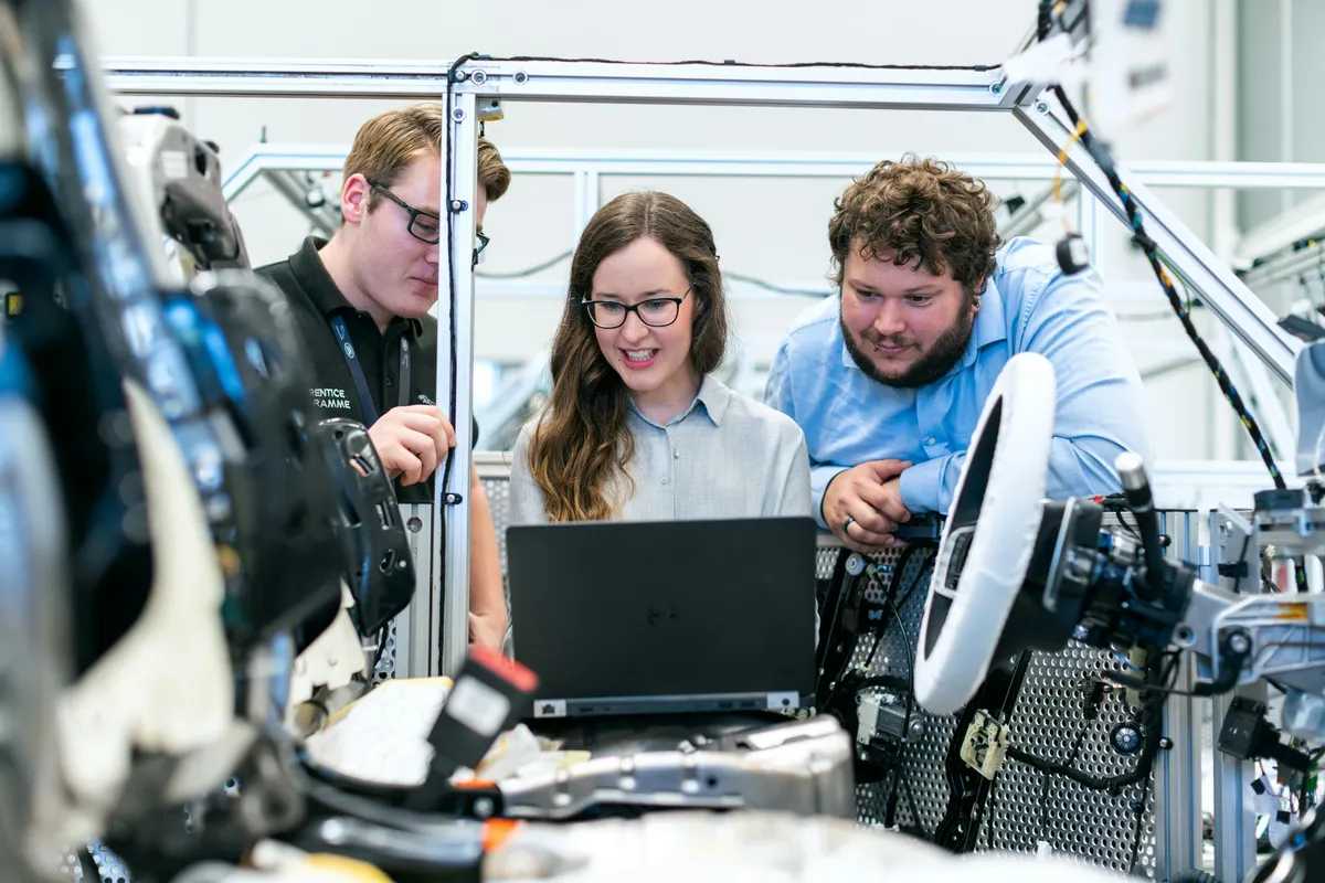 2 men and 1 woman in a manufacturing environment looking at a laptop.