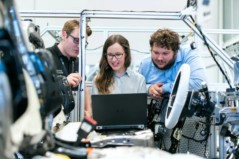 2 men and 1 woman in a manufacturing environment looking at a laptop.